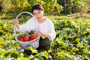 Portrait of sitting Asian woman holding basket full of harvested vegetables in her garden