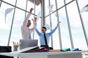 Two Asian professional businessmen celebrating their successful project by throwing document papers in the air with funny happy smiles in the daytime. Concept of achievement of a new start-up company