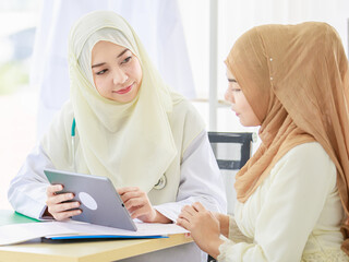 Healt concept, attractive smiling adult Muslim doctor wearing cream hijab sitting at the table and talking to the patient while using a digital tablet. Concept of healthcare services