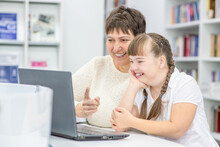 Happy Teacher And Smiling Girl With Down Syndrome Use A Laptop At Library. Education For Disabled Children Concept