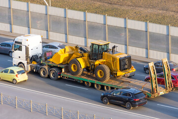 Truck with a long trailer platform for transporting heavy machinery, loaded tractor with a bucket. Highway transportation traffic jam.