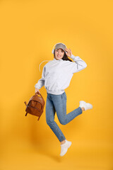 Poster - Beautiful young woman with stylish leather backpack and headphones jumping on yellow background