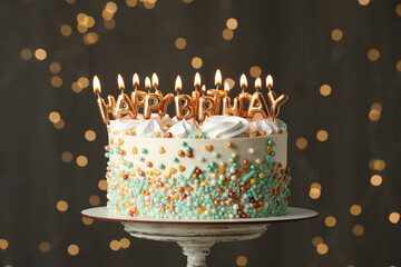 Beautiful birthday cake with burning candles on stand against festive lights