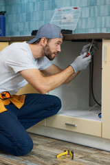 Wall Mural - Fixing leaks. Focused young repairman, professional plumber wearing tool belt crouching on the floor while fixing sink pipe in the kitchen indoors