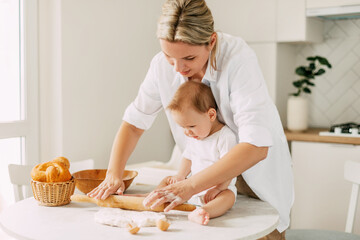 a young mother rolls out the dough on the table, her little son is sitting next to her. mom and baby