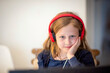 Smiling little European girl with headphones watching video lesson on computer in kitchen, happy little kid with headphones has online web class using laptop at home, home learning concept, selective