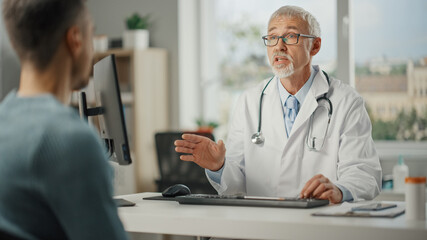 Wall Mural - Middle Aged Family Doctor is Talking with Young Male Patient During Consultation in a Health Clinic. Senior Physician in Lab Coat Sitting Behind a Computer Desk in Hospital Office.