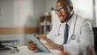 © Gorodenkoff - Happy and Smiling African American Male Doctor Wearing White Coat Working on Tablet Computer at His Office. Medical Health Care Professional Working with Test Results, Patient Treatment Planning.