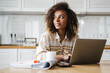 © Drobot Dean - The confident black woman typing something at the laptop and looking to the side while sitting at the table with papers in the kitchen