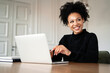 © muse studio - The freelancer works remotely on a laptop. A satisfied female designer smiles at an Afro-ethnic appearance. In the browser, working on a new work project. Curly black hair.