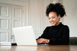 © muse studio - A satisfied female designer smiles at an Afro-ethnic appearance. A freelancer works remotely on a laptop in a browser on a new project for work. Curly black hair.