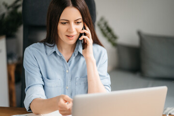 Attractive business woman sits at table in front of laptop and uses smartphone for work. Beautiful brunette girl is undergoing online training while at home. Female millennial surfing the internet