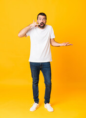 Wall Mural - Full-length shot of man with beard over isolated yellow background making phone gesture and doubting