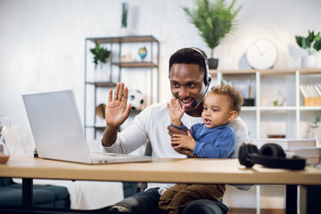 Afro american businessman with his little son on hands leading working meeting through video chat from home. Young father using modern laptop for work.