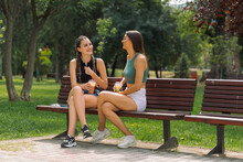 Close-up of two young girlfriends sitting on a bench in summer park chatting and having fun