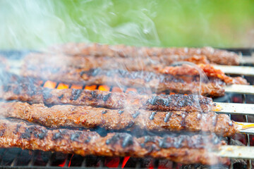 Cooking delicious Turkish Adana Kebab on a portable charcoal grill at a picnic location, Smoke and flame comes out of the sizzling meat. Koftes are lined up on flat metal skewers