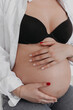 © Mikalai - Pregnant woman with hands on her belly dressed in black bra, jeans, white shirt closeup