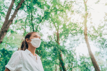 Mask wearing woman looking up to bright future in hope walking in forest. Sustainable fashion fabric in PPE, asian girl lifestyle breathing clean air.