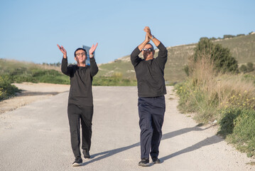 Carefree senior father and teenager son dancing with arms raised outside in a nature on a sunny summer day. Family dancing together. Young kid and elderly man having fun on a sunny summer day.