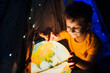 A little boy in glasses sits at night in a tent and plays with a globe.