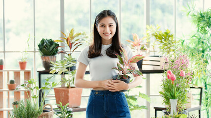 Girl long hair wear t shirt holding red Dieffenbachia tree pot smiling standing in white mirror room looking at camera. Surround by air filtering plants colorful flower spring sunlight feeling warm.