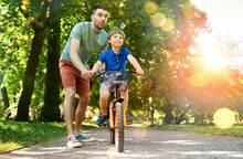 Family, Fatherhood And Leisure Concept - Happy Father Teaching Little Son To Ride Bicycle At Park