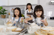 © Bangkok Click Studio - Two little cute long black fair hair Asian girl, 3 years and 7 years old, learning how to make bread and bakery with a curious and happy smiley face. Their mom standing and looking behind