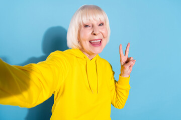 Photo of cheerful old woman good mood take selfie make v-sign hello isolated on pastel blue color background