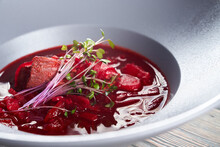 Close Up View Of Bright Red Borsch, Beet Soup With Meat And Vegetables Served With Young Green Sprouts. Tasty Hot Dish In White Plate In Restaurant. Concept Of Traditional Ukrainian Cuisine.