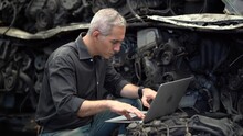 Senior Engineer Using Laptop Computer Checking List Old Used Car Engine In Machinery Parts Shop Warehouse. Old Technician Man Repair Vehicle Automobile Machine In Recycle Motor Factory