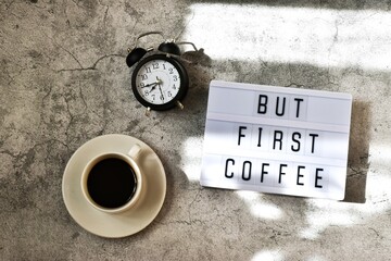 Light box with an inscription BUT FIRST COFFEE with cup coffee and alarm clock on gray concrete background. Flat lay.