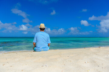 Young man sitting on the beach looking towards the horizon with blue and white stripped shirt, beach hat