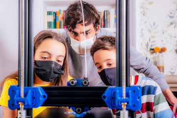 A young adult and two children wearing face masks watch how a 3D printer works.