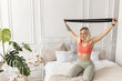 © shunevich - Happy young woman training in the morning, sitting on bed, doing exercises for the arms. Sportive blonde-hair girl stretching out, working out at home. Healthy lifestyle concept
