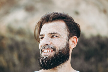 portrait of a stylish young man with a beard and a face on which the sea sand on the background of the sea shore