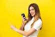 © luismolinero - Caucasian woman isolated on yellow background using mobile phone and pointing back