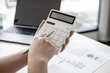 © kamiphotos - A close-up of a business finance woman's hand pressing a white calculator to calculate numbers from company financial documents. Concept of validating the accuracy of financial numbers.