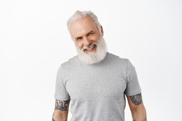 Close up of handsome hipster senior man with tattoos, tilt head, smile and look happy and friendly at camera, laughing, standing against white background