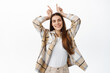 © Cookie Studio - Funny and happy adult woman playing around, showing bull devil horns gesture and smiling positive, being stubborn or determined, standing over white background