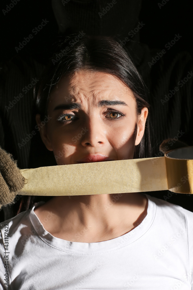 Terrorist putting sticky tape on hostage's mouth against dark background