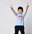 Isolated studio portrait of preschool little smiling asian boy, he wearing trendy fashion raglan with cool wolf print and sportive pants showing victory sign with two hands looking at camera