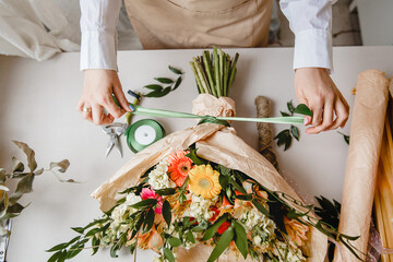 a female florist ties a green ribbon bow on a bouquet of flowers wrapped in craft paper on the deskt