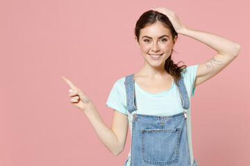 Wall Mural - Young happy surprised caucasian woman in trendy denim clothes blue t-shirt point index finger aside on workspace area mock up copy space hold face isolated on pastel pink background studio portrait