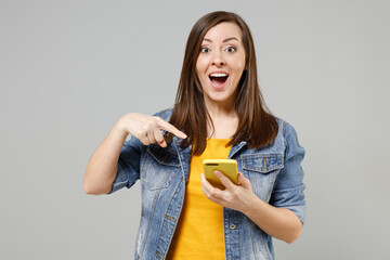 Wall Mural - Young surprised excited happy caucasian woman 20s in casual trendy denim jacket yellow t-shirt point index finger on mobile cell phone browsing chat online isolated on grey background studio portrait