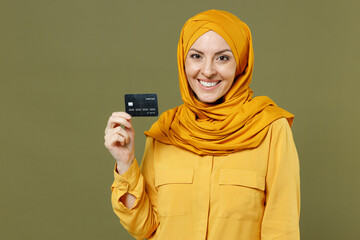 Smiling rich successful young arabian asian muslim woman in abaya hijab yellow clothes holding credit bank card isolated on olive green background studio portrait. People uae islam religious concept.