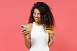 © ViDi Studio - Young happy positive african american woman 20s in casual white tank shirt hold in hand using mobile cell phone credit bank card pay online do shopping purchasing isolated on pink color background