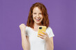© ViDi Studio - Young smiling happy satisfied fun redhead woman in white basic casual t-shirt holding using mobile cell phone do winner gesture clench fist isolated on dark violet color background studio portrait