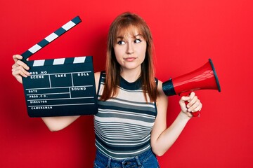 Sticker - Redhead young woman holding video film clapboard and megaphone smiling looking to the side and staring away thinking.