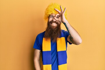 Canvas Print - Redhead man with long beard football hooligan cheering game wearing funny wig doing ok gesture with hand smiling, eye looking through fingers with happy face.