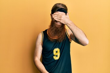 Poster - Redhead man with long beard wearing basketball uniform smiling and laughing with hand on face covering eyes for surprise. blind concept.